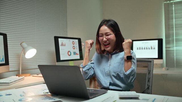 Excited asian business analyst working overtime on her laptop, celebrating a professional victory and goal achievement with a joyful gesture while sitting at her desk in the office at night