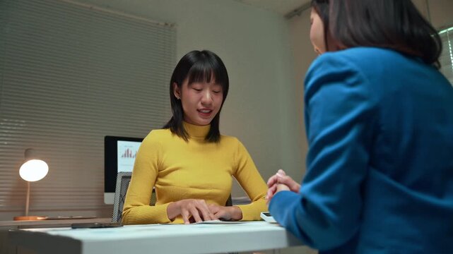 Two young Asian businesswomen discussing a project, reviewing documents and brainstorming ideas together during a late-night meeting in a modern office, showing teamwork and dedication