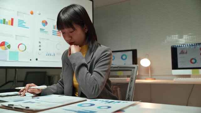 Pensive young asian businesswoman analyzing financial market reports and studying business statistics charts on papers, working overtime at her desk in a modern office late at night