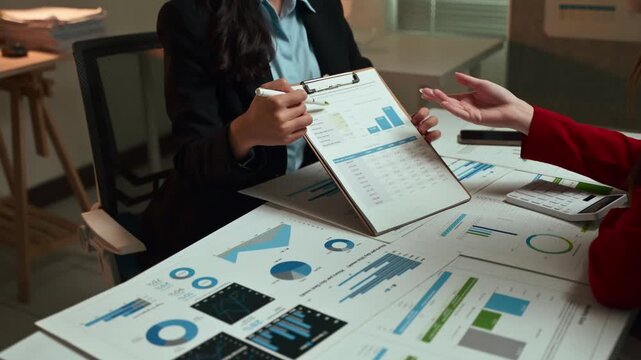 Two businesswomen discussing financial reports and analyzing statistical data with graphs and charts during a late-night meeting, collaborating on a new business project strategy