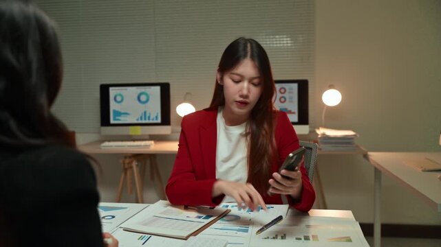 Stressed Asian businesswoman shouting and arguing with a colleague during a tense late-night meeting, using a calculator on her phone to discuss financial figures and company expenses
