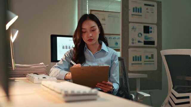 Diligent asian businesswoman working late at night in the office, carefully examining documents on a clipboard and interacting with her computer screen to complete her tasks before the deadline