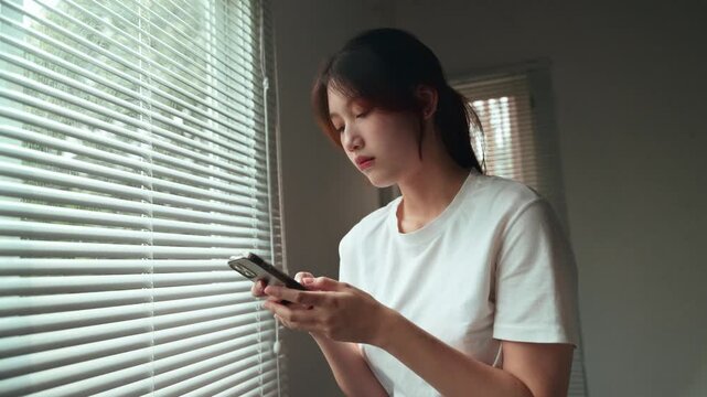 Young Asian woman with a concerned expression using her mobile phone while standing near a window with blinds, looking thoughtful and worried after reading a message in her home
