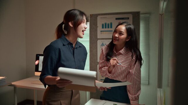 Young Asian businesswomen collaborating on a project, discussing financial charts and brainstorming ideas while working overtime in a modern office to meet a critical deadline