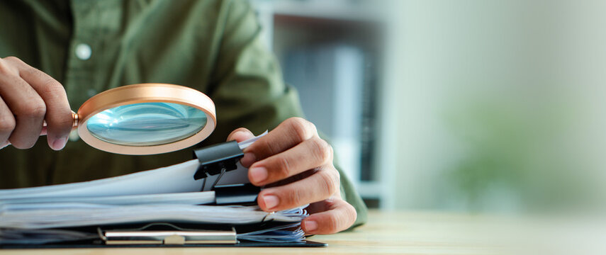 Auditor inspects business documents with a magnifying glass on desk. Compliance review, financial investigation, accounting verification, paperwork audit, risk assessment, and fraud detection concept.