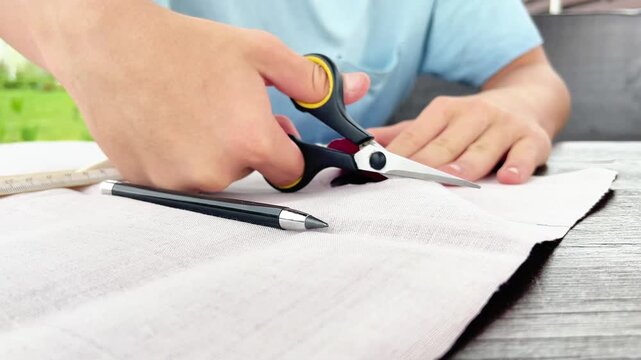 Close-up of a person using sharp scissors to cut along a pencil line on light linen fabric during a DIY craft project at a wooden table.