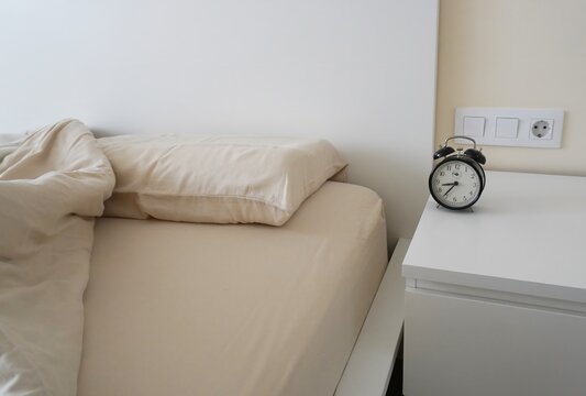 A morning scene with beige linens and a black alarm clock on a white bedside table. An empty unmade bed with a black alarm clock.