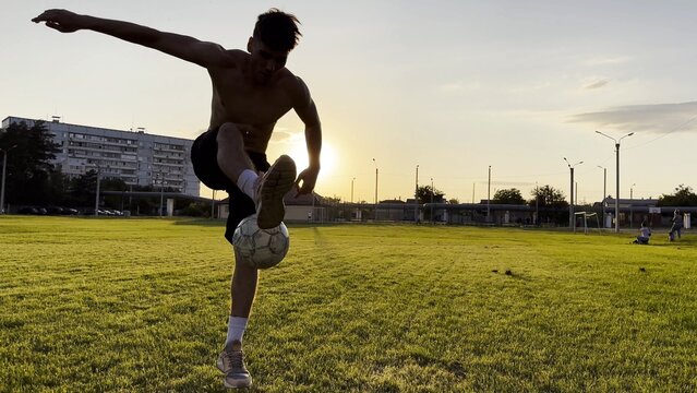 Young man juggling soccer ball on stadium at sunset. Professional footballer kicking ball at green field. Sportsman practicing tricks at meadow with sunlight at background. Freestyle football