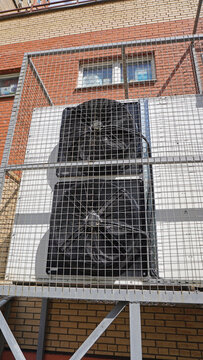 Industrial fans are installed in a lattice casing on the front panel. Vertical installation, metal frame, window above, brick wall, daylight.