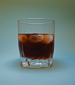 A glass with a dark drink and ice on a blue background. Condensation drops, clear glass, studio light. Background light on a cocktail glass. Whiskey with ice. Cognac with ice.
