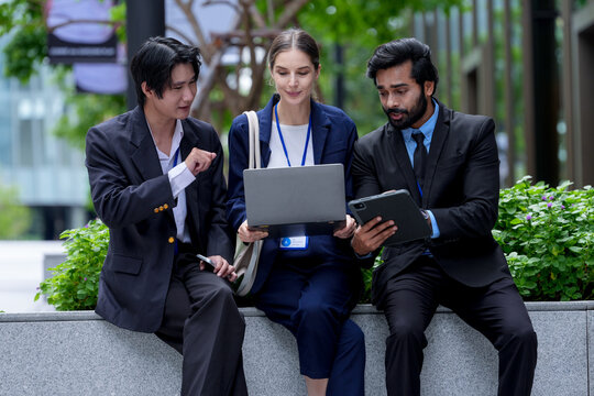 Three young business professionals wearing conference badges review a document together on the laptop screen under fresh trees in the public realm of a smart city (frame 99)