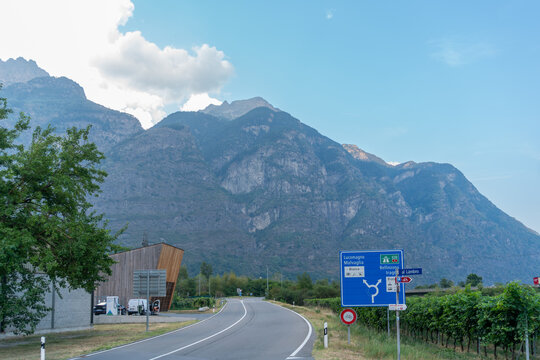 Scenic road runs through a vineyard beneath massive rocky mountains in Switzerland. Directional signs show routes toward Lucomagno, Malvaglia, Biasca and Bellinzona, creating an authentic alpine road 