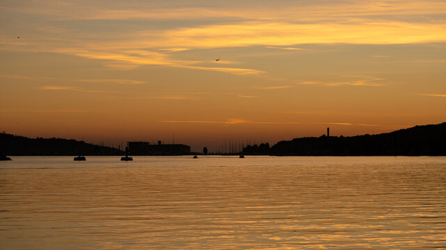 Warm golden sunset over the calm waters of Pula harbour in Croatia, with distant industrial waterfront structures, hills and silhouettes reflected across the Adriatic Sea.