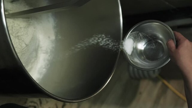 Vertical video. Culinary process. Dough preparation. Baker adding measured ingredients in industrial mixer. Hand pouring salt in flour.
