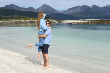 Happy young man lifting his girlfriend on a white sand beach of Sommaroy in Norway. Loving couple embracing and having fun on a scenic arctic coast with mountains. © vaitekune