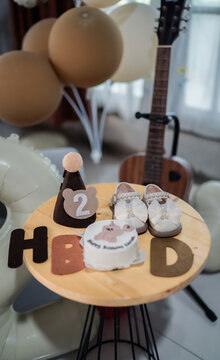 Cute 2nd birthday party setup with small cake, baby shoes, party hat, HBD letters, and balloons in brown and cream tones.