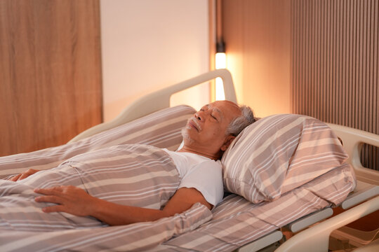 Elderly Asian man sleeping peacefully in an adjustable nursing care bed under warm lamplight in a cozy bedroom of a senior home, showing gentle rest and patient comfort (0880)