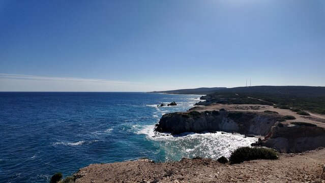 Harsh cliffs rise above the deep blue sea, where waves crash against rocks and the coastline stretches into the distance under a clear sunny sky.