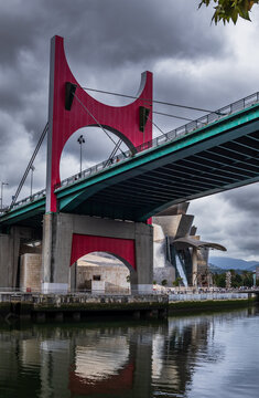 Vista en contrapicado de la llamativa estructura roja del puente de La Salve sobre la r&iacute;a de Bilbao, con el Museo Guggenheim al fondo bajo un cielo nubla