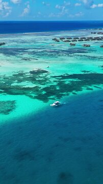High angle aerial shot of a tropical island and boat in the Maldives, featuring transparent ocean water under a blue sky with rich blue and green accents in the setting. Vertical video