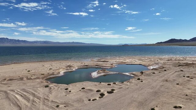 puddles on dry sandy beach near lake shore with blue sky and mountains in background