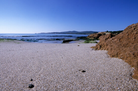 a sandy beach with reddish-brown rock formations and calm blue sea under a clear sky.. Spiaggia delle Bombarde, sullo sfondo Capo Galera. Alghero. SS, Sardegna. Italia
