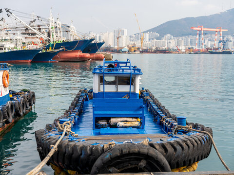 View of a blue tugboat moored in the harbor with large fishing vessels, industrial cranes, and the city skyline in the background in Busan, Busan, South Korea.