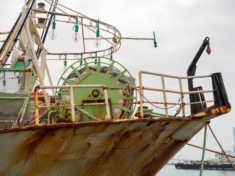 View of the weathered bow of a fishing boat featuring a large green net winch, metal railings, and ropes docked in the harbor of Busan, Busan, South Korea.