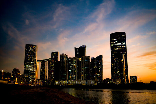 View of the illuminated Al Reem Island city skyline at dusk with modern skyscrapers reflecting against the calm water in Abu Dhabi, Abu Dhabi, United Arab Emirates.