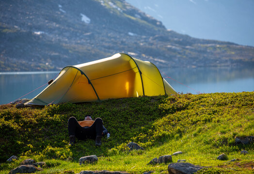 View of a person resting in the shade on the grass beside a yellow tent near a tranquil lake with mountain peaks in the background in Fjord, More og Romsdal, Norway.