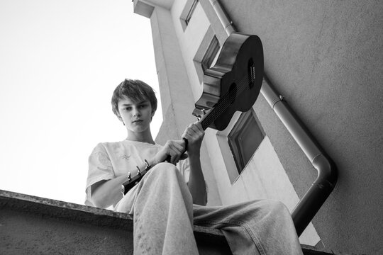 teenager sitting on rooftop ledge playing acoustic guitar, building wall with window and drain pipe behind, bright sky, low angle view, music hobby, leisure and free time outdoors in daylight