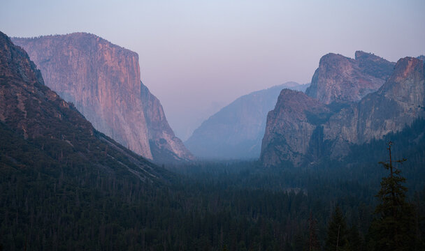 View of El Capitan and the granite cliffs of Yosemite Valley at dusk with hazy sky and dense forest in Yosemite Valley, California, United States.
