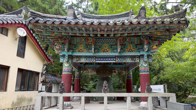 View of a traditional Korean temple bell pavilion with intricate dancheong painting and a large bronze bell, surrounded by lush trees on Jangsan Mountain, Busan, South Korea.