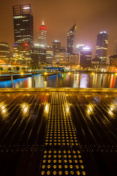View of the illuminated Elizabeth Quay and the Perth skyline reflected in the water at night, featuring a wooden boardwalk with tactile paving in Perth, Western Australia, Australia.