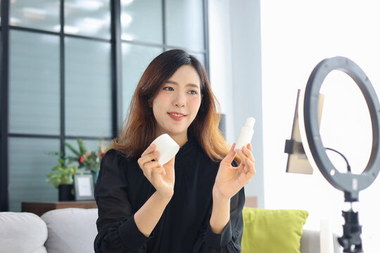 Asian woman filming beauty vlog tutorial with ring light, reviewing cosmetics cream moisturizer or lotion.