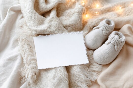 Baby announcement mockup with blank card and knitted booties. Cozy flat lay composition on a beige blanket with fairy lights. Copy space for text