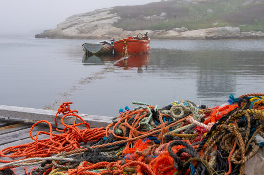 Colorful fishing ropes lie on a wooden wharf overlooking traditional boats moored in a foggy harbor at Peggy&rsquo;s Cove, NS.