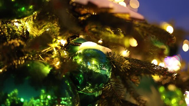 close-up of a shiny green glass Christmas ornament hanging on a snow-dusted pine branch, reflecting warm string lights and a glimpse of the winter scene around it