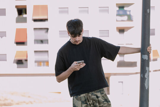 Young man using smartphone outdoors in the city, looking down at his mobile phone with a focused expression while leaning on a street pole in front of an apartment building on a sunny day