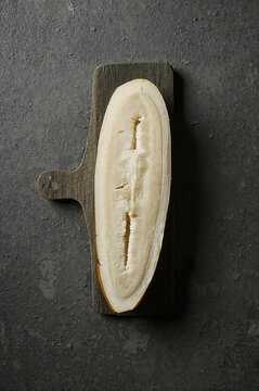 A vertical slice of a pale yellow vegetable, possibly a cucumber or squash, resting on a small, dark wooden cutting board against a gray surface