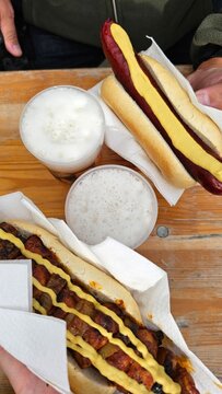 Zwei Hotdogs mit W&uuml;rstchen Senf Ketchup und R&ouml;stzwiebeln auf einem Holztisch mit Getr&auml;nken als beliebtes Streetfood