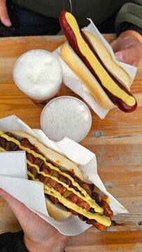 Zwei Hotdogs mit W&uuml;rstchen Senf Ketchup und R&ouml;stzwiebeln auf einem Holztisch mit Getr&auml;nken als beliebtes Streetfood