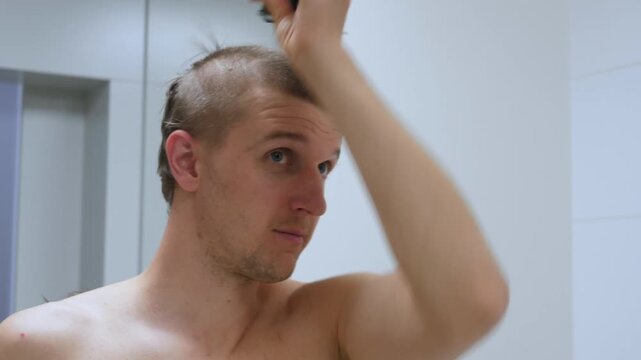 Young man trims his hair with electric clipper in bathroom mirror, focused and calm. Shirtless grooming routine continues as he shapes his buzz cut with steady hand near white tiles.