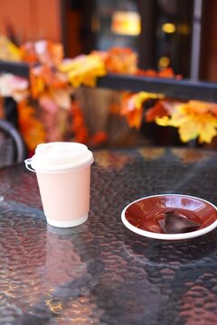 Takeaway coffee cup with chocolate candy on glass table of street cafe with autumn leaves decor. Self-care, cozy fall break and Halloween season concept. Warm seasonal atmosphere outdoors.
