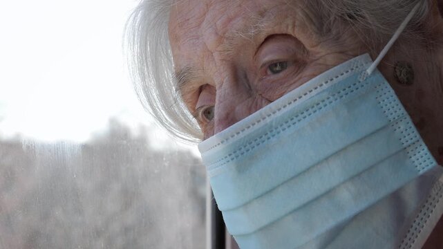 Elderly woman wearing protective face mask looking sadly out window at home. Old female expressing loneliness and isolation during virus pandemic quarantine, feeling vulnerable and at high risk