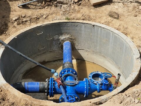 Underground utility infrastructure featuring a circular concrete manhole or access chamber partially filled with muddy water. Inside, large blue painted ductile iron water pipes with flanged