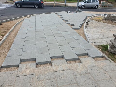 Construction site showing installation of gray concrete paving stones for a pedestrian walkway or plaza. Rectangular slabs are laid in a staggered pattern on a sand base, with some stones still