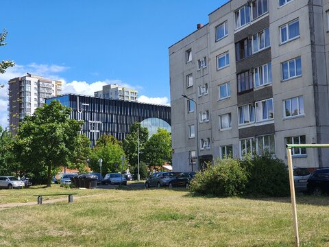 An urban scene contrasting modern and older architecture under a bright blue sky. In the foreground, a grassy area with scattered trees and a wooden bench borders a parking lot with several cars. To