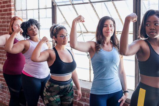 Diverse women flexing biceps in a bright studio, confident fitness pose