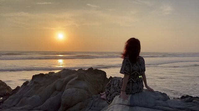 Static shot of woman with red hair sitting and leaning on coastal rocks, relaxing during a golden ocean sunset